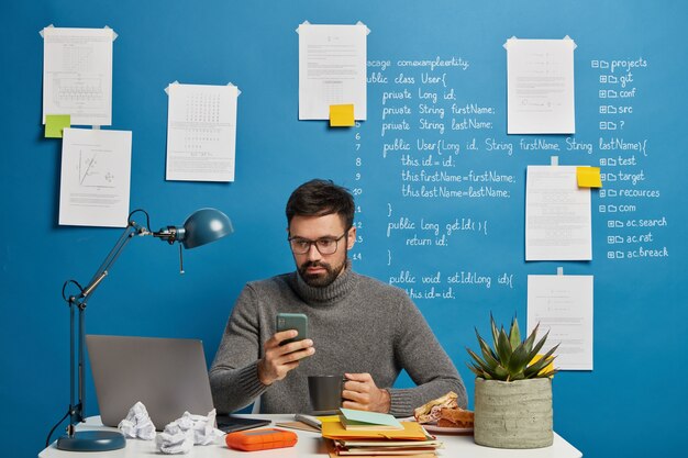 IT-Profi arbeitet am Startprojekt, aktualisiert Software und Datenbank auf dem Handy, trinkt heißes Getränk und sitzt mit schriftlichen Informationen am Desktop an der blauen Wand.