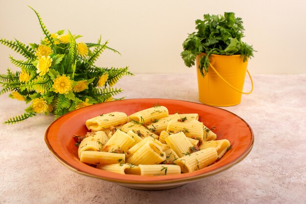 Italienische Nudeln mit Vorderansicht, lecker gekocht mit getrocknetem Grün und gesalzenem runden orangefarbenen Teller mit Blume auf rosa Schreibtisch