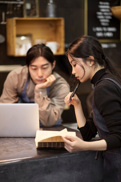 Japanische Frau liest aus dem Notizbuch, während der Japaner in einem Restaurant an seinem Laptop arbeitet