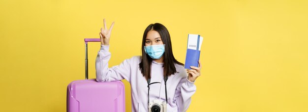 Japanisches Mädchen in medizinischer Maske, das ihren Pass und ihre Flugtickets zeigt Friedenszeichen posiert