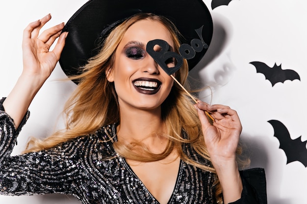 Jocund blondes Mädchen genießt Halloween-Party. Blithesome weibliches Modell im Hut, das mit Fledermäusen an der Wand aufwirft.