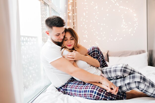 Jocund dunkelhaariges Mädchen, das Hände mit Ehemann beim Aufstellen im Bett hält. Innenfoto des kühlenden europäischen Liebespaares.