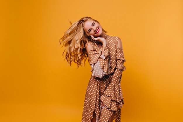 Jocund lachendes Mädchen im Retro-Kleid, das auf gelber Wand steht. Charmante elegante Frau, die Fotoshooting genießt.