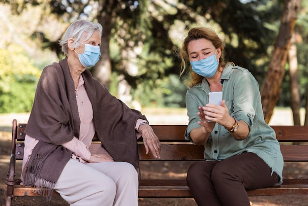 Jüngere frau mit medizinischer maske, die ältere frau auf bank etwas auf smartphone zeigt