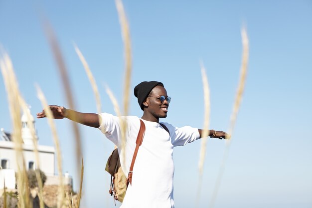 Jugend, Glück, Freiheit und Reisen. Fröhlicher junger afroamerikanischer Mannreisender, der Hut und Sonnenbrille trägt, die Arme ausbreitet und glücklich lächelt, sich entspannt und im Einklang mit der Natur fühlt