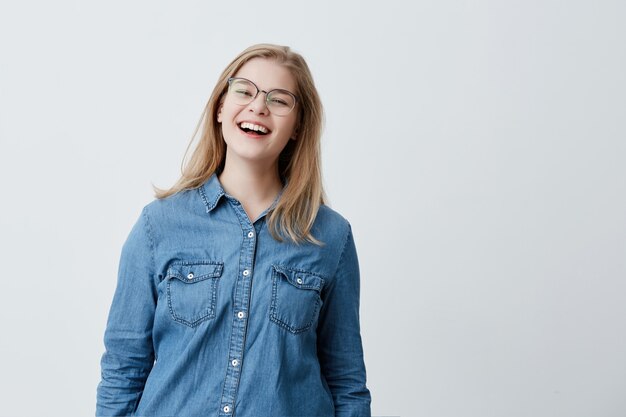 Jugend-, Lebensstil- und Bildungskonzept. Hübsches Studentenmädchen mit blonden Haaren und breitem Lächeln, lachend, schauend, während sie sich zu Hause ausruhen, trendige Brillen und Jeanshemd tragen