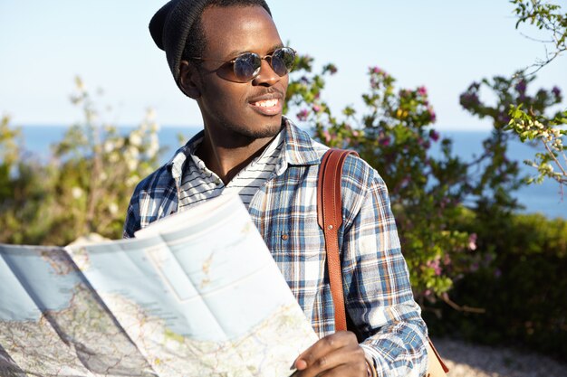 Jugend, Lebensstil und Reisen. Dunkelhäutiger männlicher Reisender in Sonnenbrille und Rucksack mit Straßenkarte, die seine Reise genießt