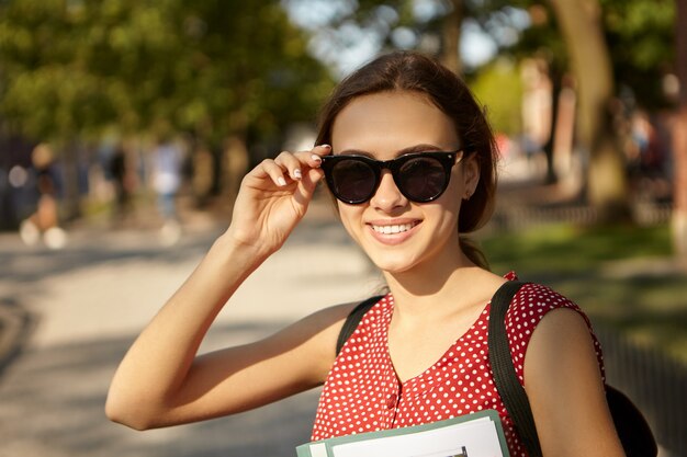 Jugend-, Sommer-, Stil- und Glückskonzept. Fröhliches modisches hübsches Mädchen mit schlankem Körper und niedlichem glücklichem Lächeln, das Hand auf ihrer schwarzen Sonnenbrille hält, schöne Zeit im Freien verbringend