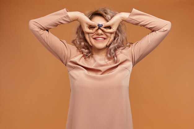 Jugend, Spaß und Freude Konzept. Schönes lustiges Hipster-Mädchen mit lockigem rosa Haar und Nasenring, die Schutzbrille mit ihren Fingern bilden, Daumen und Zeigefinger verbindend