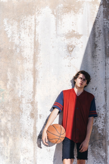 Jugendbasketballspieler, der auf Wand am sonnigen Tag sich lehnt