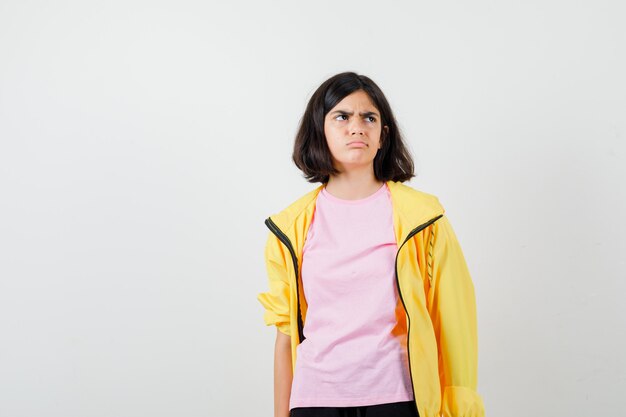 Jugendlich Mädchen, das im gelben Trainingsanzug, im T-Shirt nach oben schaut und unzufrieden aussieht, Vorderansicht.