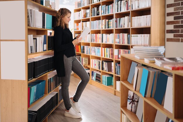 Jugendlich Mädchen unter einem Stapel Bücher. Ein junges Mädchen liest ein Buch mit Regalen im Hintergrund. Sie ist von Bücherstapeln umgeben. Tag buchen.