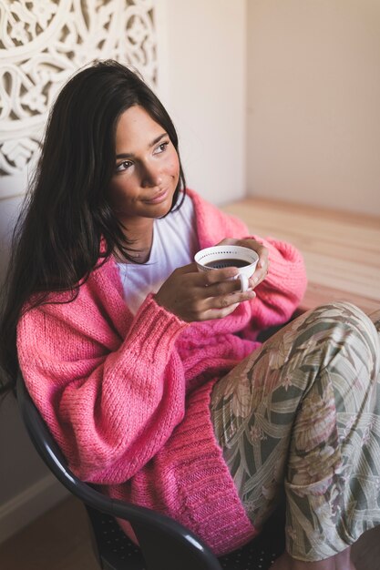 Jugendliche, die auf dem Stuhl trägt die rosa Strickjacke hält Tasse Kaffee sitzt