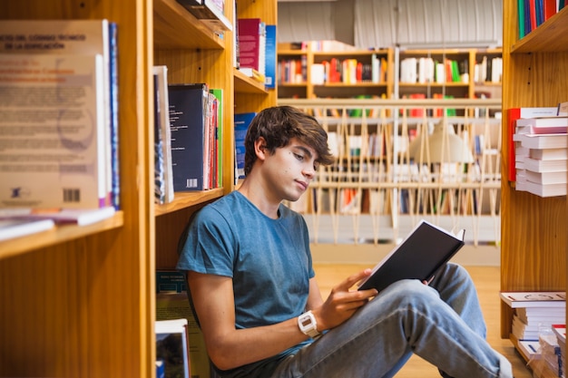 Jugendlicher, der auf Fußboden zwischen Bücherregalen liest