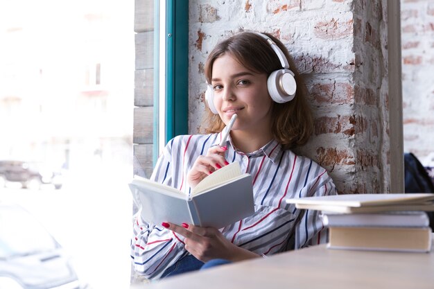 Jugendlichmädchen in den Kopfhörern, die mit offenem Buch sitzen und Kamera betrachten