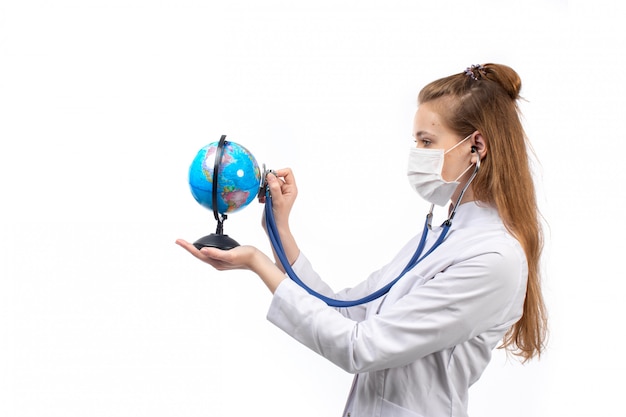 Kostenloses Foto junge ärztin im weißen medizinischen anzug mit stethoskop in der weißen schutzmaske, die den kleinen globus auf dem weiß hört