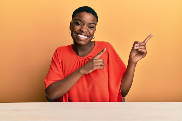 Junge afrikanisch-amerikanische Frau in Freizeitkleidung sitzt lächelnd auf dem Tisch und schaut in die Kamera, die mit zwei Händen und Fingern zur Seite zeigt.