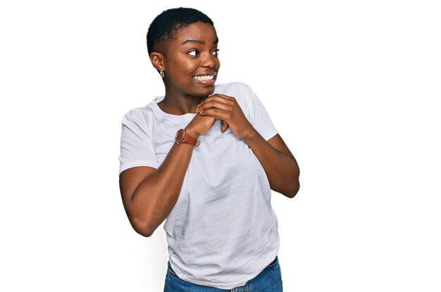 Junge afrikanisch-amerikanische Frau in lässigem weißem T-Shirt lacht nervös und aufgeregt mit den Händen am Kinn und schaut zur Seite