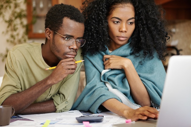 Junge afrikanische Frau und Ehemann mit vielen Schulden, die Papierkram zusammen machen, Ausgaben analysieren, Familienbudget planen und Rechnungen berechnen, am Küchentisch mit Laptop, Taschenrechner und Papieren sitzen