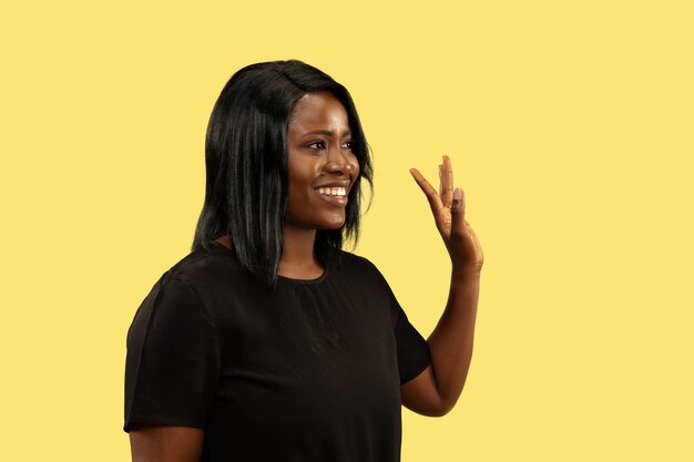 Junge Afroamerikanerfrau lokalisiert auf gelbem Studiohintergrund, Gesichtsausdruck. Schönes weibliches halblanges Porträt. Konzept menschlicher Emotionen, Gesichtsausdruck. Zeige das Zeichen des Tschüss.