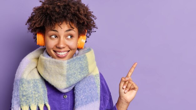 Junge Afroamerikanerin mit Kopfhörern, trägt einen warmen Schal um den Hals, fühlt sich glücklich und zeigt auf den Kopierraum