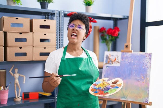 Junge afroamerikanische Frau, die im Kunststudio auf Leinwand zeichnet, wütend und wütend schreiend, frustriert und wütend schreiend vor Wut, Wut und aggressivem Konzept