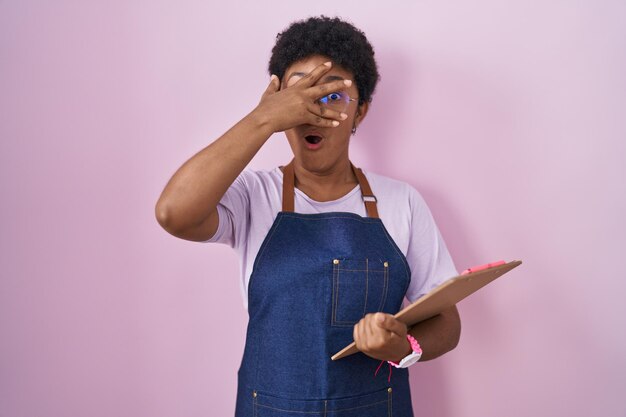 Junge afroamerikanische Frau trägt eine professionelle Kellnerschürze, hält ein Klemmbrett in der Hand und blickt schockiert über Gesicht und Augen, während die Hand mit verlegenem Gesichtsausdruck durch die Finger blickt
