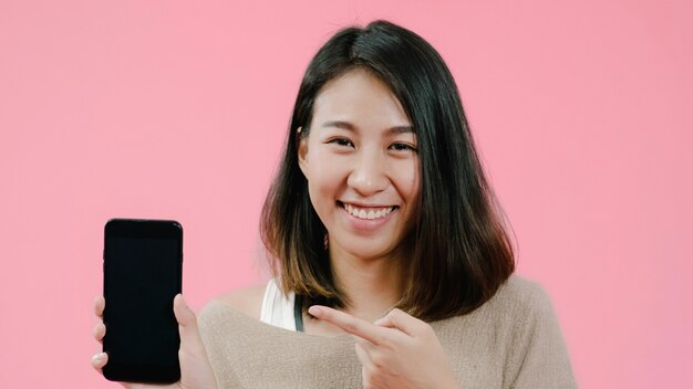 Junge Asiatin, die den Smartphone überprüft das Social Media glaubt dem glücklichen Lächeln in der Freizeitbekleidung über rosa Hintergrund verwendet