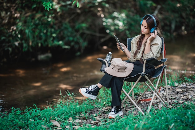 Junge asiatin sitzt auf einem stuhl in der nähe des stroms und hört glücklich musik vom tablet mit drahtlosen kopfhörern, während sie im wald campt