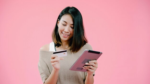 Junge Asiatin, welche die Tablette kauft Online-Shopping mit Kreditkarte fühlt sich glücklich lächelnd in der Freizeitbekleidung über rosa Hintergrundatelieraufnahme. Glückliche lächelnde entzückende frohe Frau freut sich Erfolg.