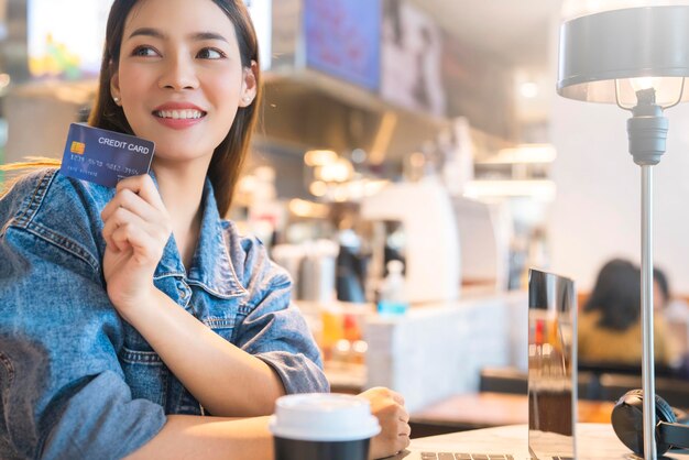 Junge asiatische attraktive weibliche reisende hand halten kreditkarte für online-einkäufe mit laptop im café-restaurant-geschäftsideenkonzept