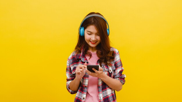 Junge asiatische Dame, die drahtlose Kopfhörer trägt, die Musik vom Smartphone mit fröhlichem Ausdruck in der Freizeitkleidung hören und Kamera über gelber Wand betrachten. Gesichtsausdruck Konzept.