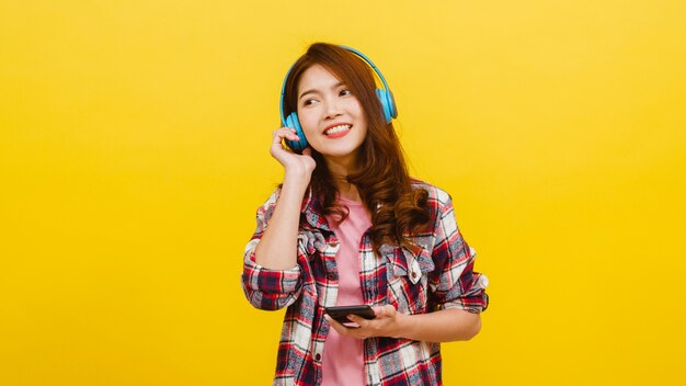 Junge asiatische Dame, die drahtlose Kopfhörer trägt, die Musik vom Smartphone mit fröhlichem Ausdruck in der Freizeitkleidung hören und Kamera über gelber Wand betrachten. Gesichtsausdruck Konzept.