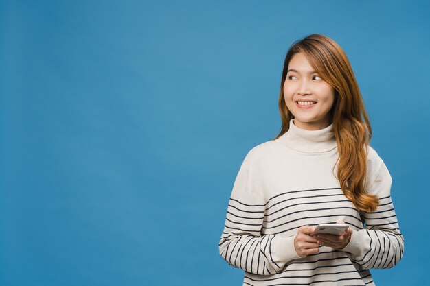 Junge asiatische Dame, die Telefon mit positivem Ausdruck benutzt, breit lächelt, in Freizeitkleidung gekleidet ist, sich glücklich fühlt und isoliert auf blauer Wand steht