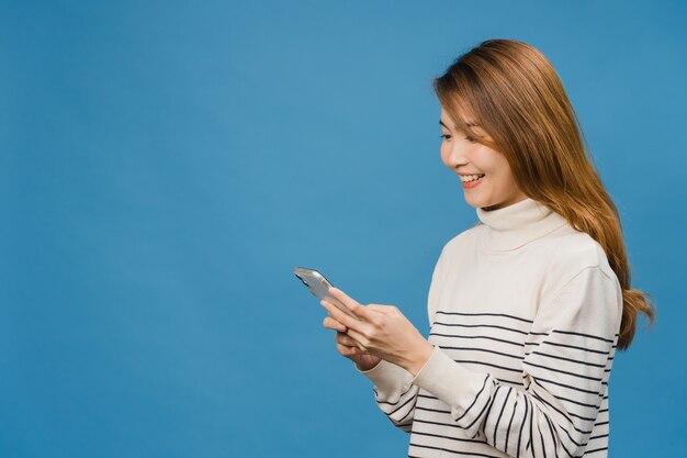 Junge asiatische Dame, die Telefon mit positivem Ausdruck benutzt, breit lächelt, in Freizeitkleidung gekleidet ist, sich glücklich fühlt und isoliert auf blauer Wand steht