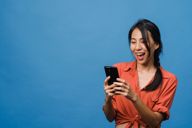 Junge asiatische Dame, die Telefon mit positivem Ausdruck benutzt, breit lächelt, in Freizeitkleidung gekleidet ist und sich glücklich fühlt und isoliert auf blauer Wand steht. Glückliche entzückende frohe Frau freut sich über Erfolg.
