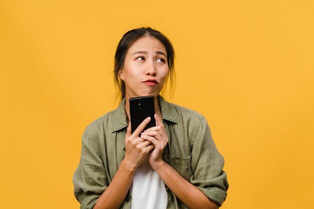 Junge asiatische Dame, die Telefon mit positivem Ausdruck benutzt, breit lächelt, in Freizeitkleidung gekleidet ist und sich glücklich fühlt und isoliert auf gelber Wand steht. Glückliche entzückende frohe Frau freut sich über Erfolg.