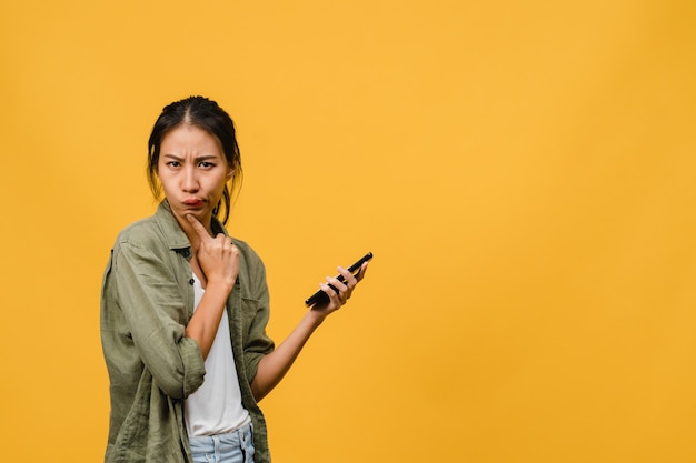 Junge asiatische Dame, die Telefon mit positivem Ausdruck benutzt, gekleidet in legerem Tuch auf gelber Wand