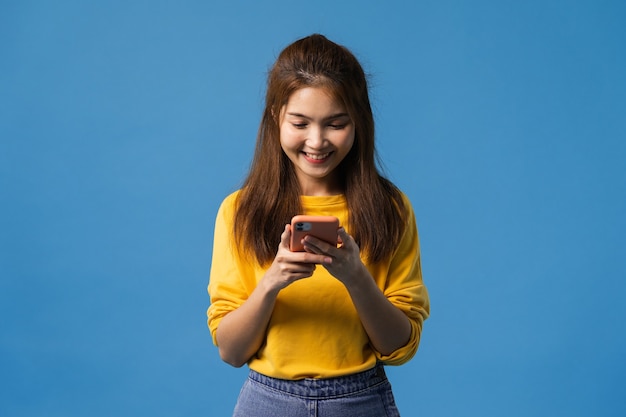 Junge asiatische Dame, die Telefon mit positivem Ausdruck verwendet, lächelt breit, gekleidet in Freizeitkleidung, die Glück fühlt und isoliert auf blauem Hintergrund steht. Glückliche entzückende frohe Frau freut sich über Erfolg.