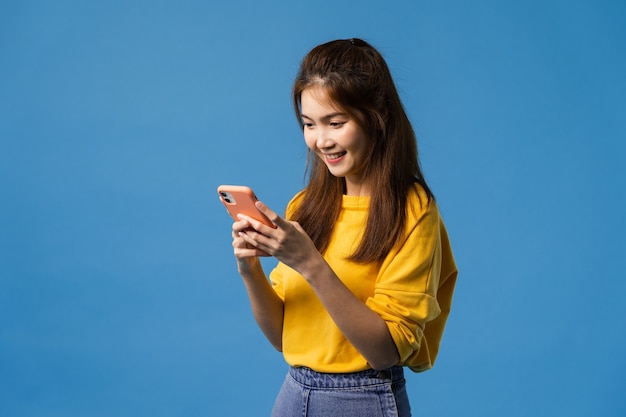 Junge asiatische Dame, die Telefon mit positivem Ausdruck verwendet, lächelt breit, gekleidet in Freizeitkleidung, die Glück fühlt und isoliert auf blauem Hintergrund steht. Glückliche entzückende frohe Frau freut sich über Erfolg.