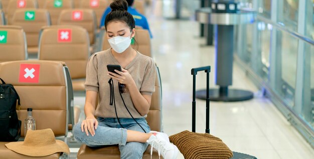 Junge asiatische erwachsene reisende mit gesichtsmaske hand halten smartphone sitzend mit sozialer distanzierung warten auf den flugplan in der lobby am flughafenterminal sicherheitskonzept für reiseideen