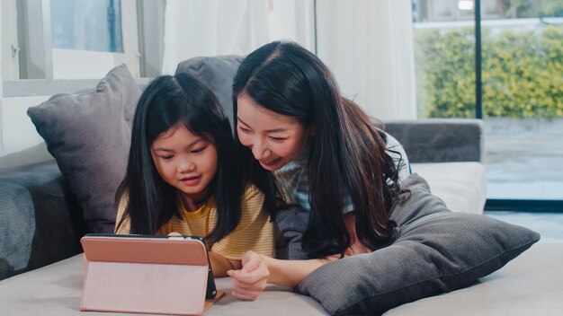 Junge asiatische Familie und Tochter glücklich, Tablette zu Hause verwendend. Japanische Mutter entspannen sich mit dem aufpassenden Film des kleinen Mädchens, der auf Sofa im Wohnzimmer am Haus liegt. Lustige Mutter und schönes Kind haben Spaß.