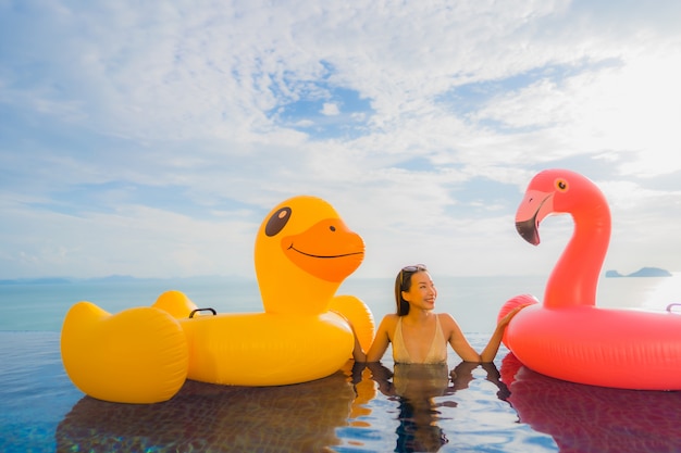 Junge asiatische Frau des Porträts auf gelber Ente des aufblasbaren Schwimmers und rosa Flamingo um Swimmingpool im Freien im Hotel und im Erholungsort