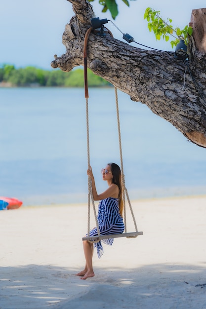 Junge asiatische Frau des Porträts, die auf Schwingenseil und Meer um Strandseeozean-Kokosnusspalme sitzt