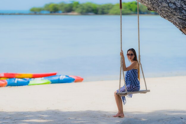 Junge asiatische Frau des Porträts, die auf Schwingenseil und Meer um Strandseeozean-Kokosnusspalme sitzt