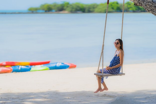 Junge asiatische Frau des Porträts, die auf Schwingenseil und Meer um Strandseeozean-Kokosnusspalme sitzt