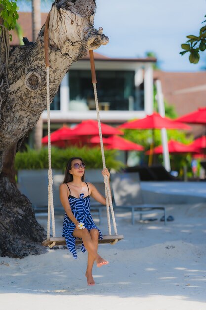 Junge asiatische Frau des Porträts, die auf Schwingenseil und Meer um Strandseeozean-Kokosnusspalme sitzt