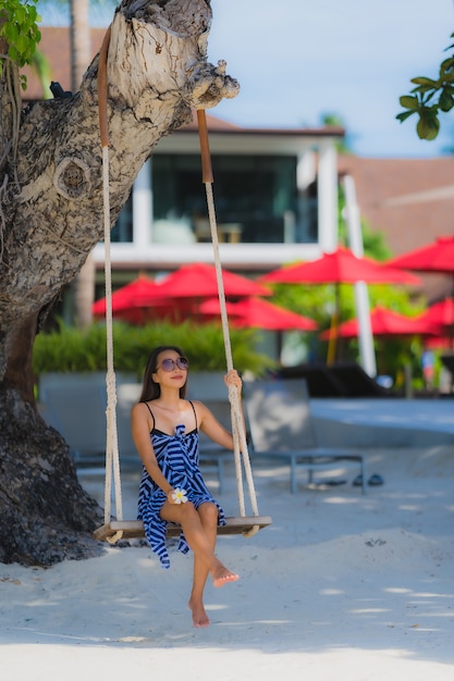 Junge asiatische Frau des Porträts, die auf Schwingenseil und Meer um Strandseeozean-Kokosnusspalme sitzt