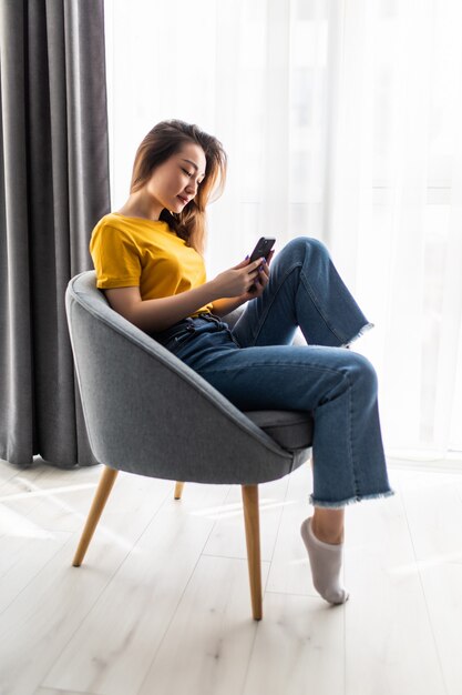 Junge asiatische Frau des Porträts, die Handy benutzt, sitzt auf Stuhl im Wohnzimmerinnenraum