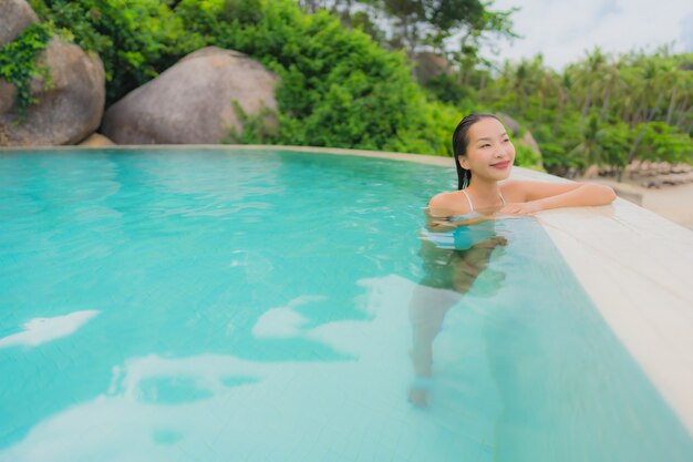 Junge asiatische Frau des Porträts entspannen sich das Lächeln, das um Swimmingpool im Freien im Hotelerholungsort mit Seeozeanblick glücklich ist